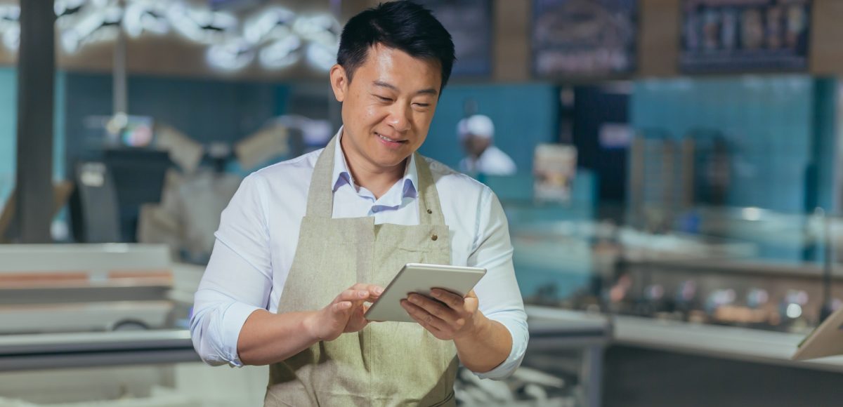 Asian grocery store manager salesman in apron using digital tablet counting goods in supermarket